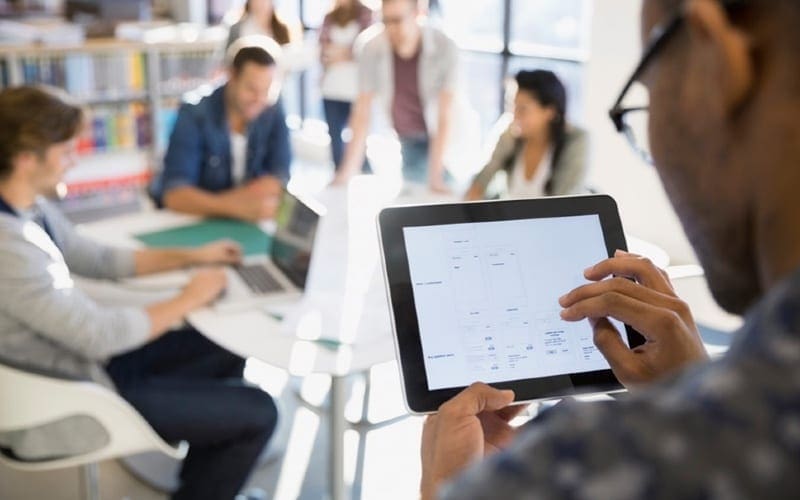 Un homme utilise une tablette dans un bureau collaboratif pour consulter des plans ou un flux de travail, symbolisant l'utilisation de solutions logicielles pour la gestion des documents et l'impression professionnelle.